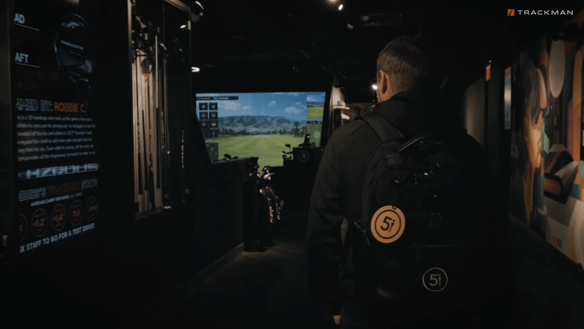 A person with a backpack walks toward a golf simulator screen in a dimly lit room.