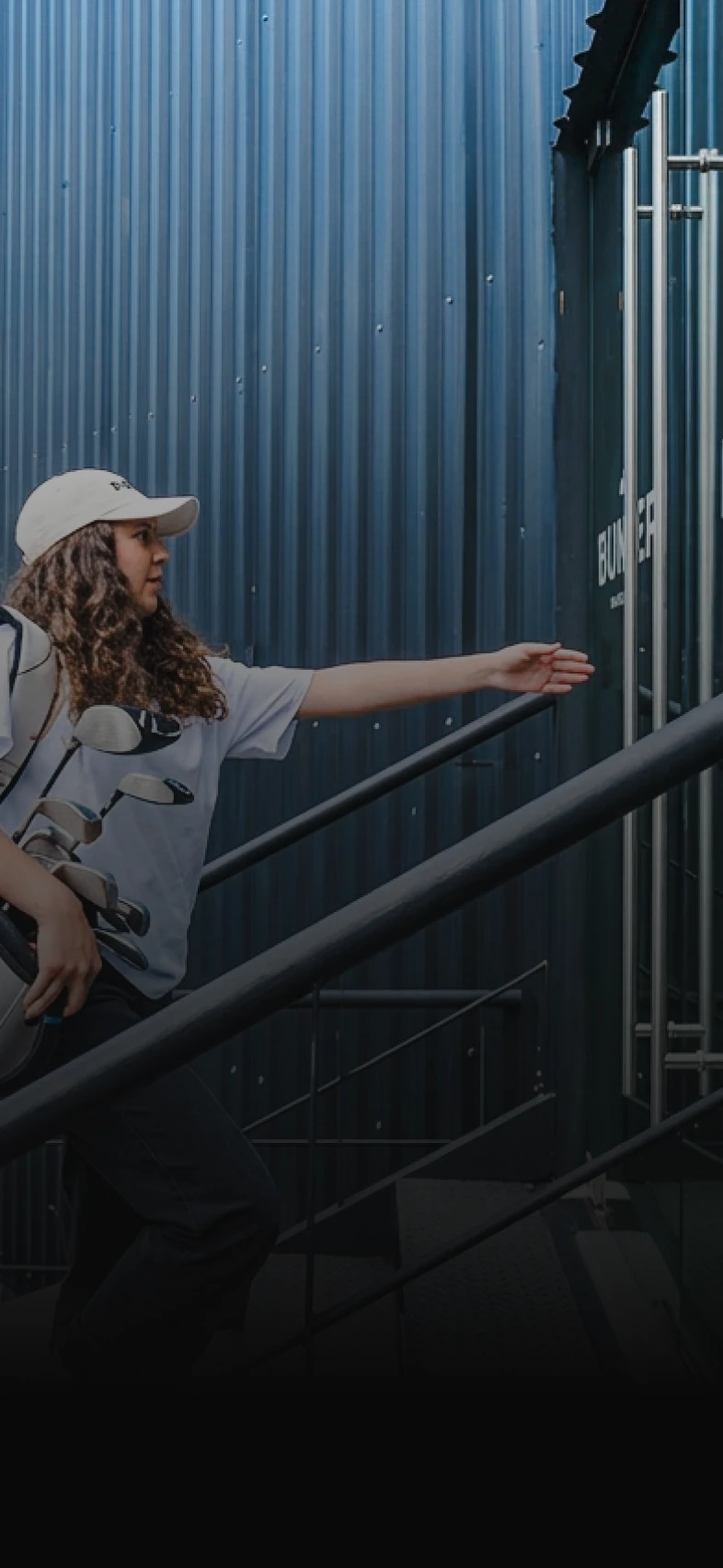 A person wearing a cap carries golf clubs while reaching for a door labeled "Bunker" on a staircase beside a corrugated blue wall.